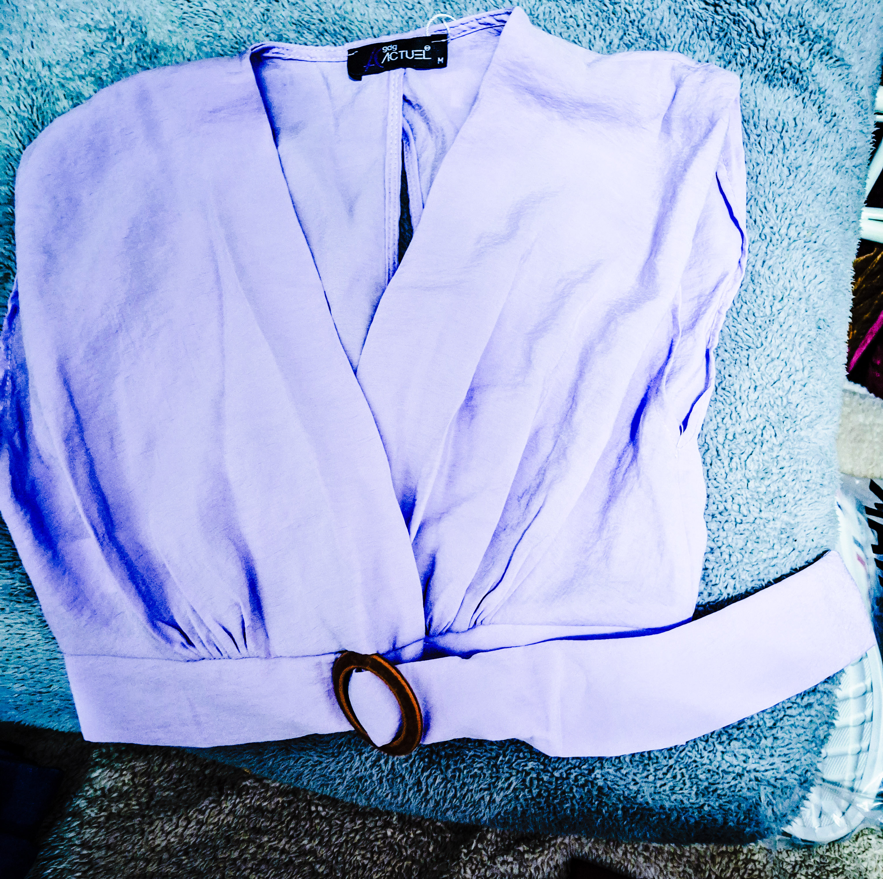 A lavender women's top with a deep v-neckline and a large circular belt buckle, laid flat on a textured blue background. Different colours and sizes. price £4.40